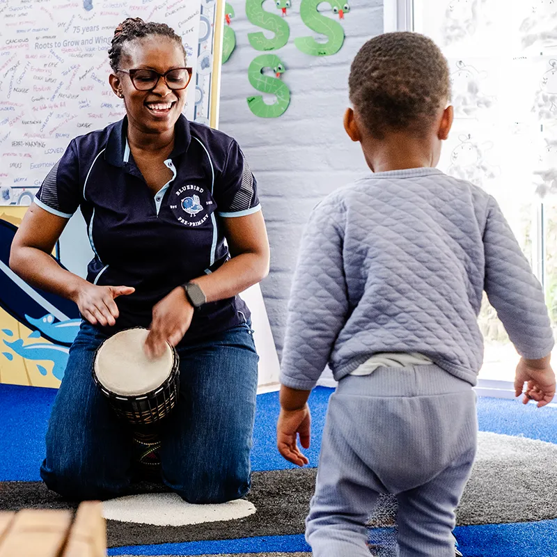 Music and movement activities encourage children to develop rhythm, coordination, listening skills and self-expression while enjoying the joy of movement and sound. Teacher playing a drum while a young child participates in a music and movement activity at Bluebird Pre-Primary.