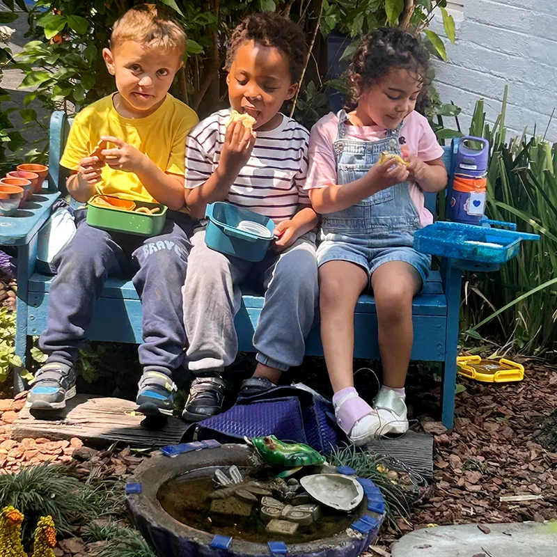 The Friendship Garden is a place for connection, conversation, and shared experiences. Whether enjoying a snack together, creating stories, or simply spending time with friends, children learn the value of kindness, belonging, and friendship. Three preschool children sit together on a blue bench in the Friendship Garden, enjoying a snack beside a small pond surrounded by plants and natural play elements.