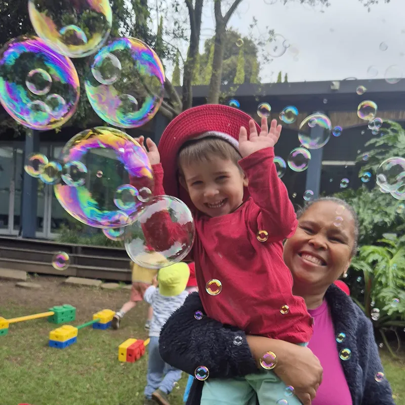 Moments of joy, connection, and wonder are part of every Bluebird day. Through play, laughter, and caring relationships, children build confidence, create happy memories, and develop a strong sense of belonging. A smiling preschool child wearing a red sun hat plays with bubbles while being held by a teacher outdoors, surrounded by colourful play equipment and a joyful garden setting.