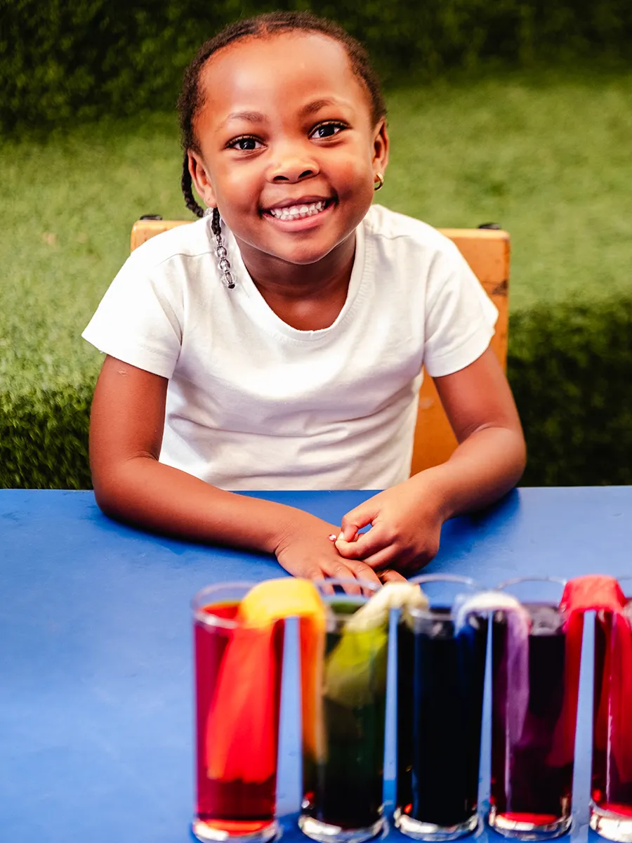 Learning through curiosity, creativity and play. A smiling Bluebird Pre-Primary learner seated at an outdoor table with colourful paint-filled glasses arranged in front of her.