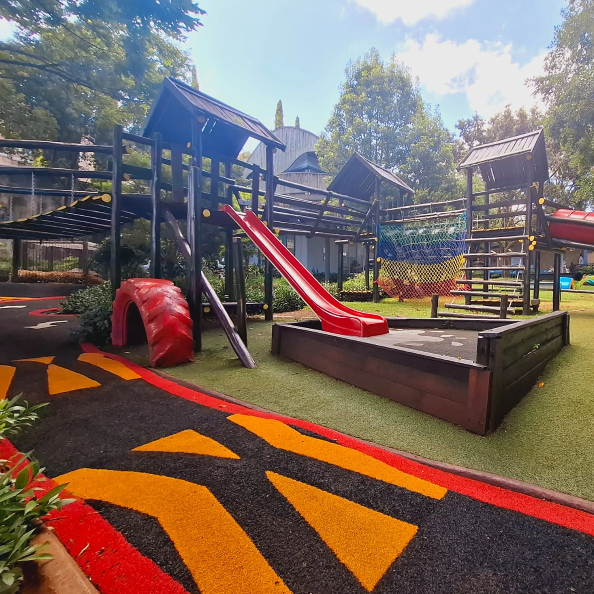 A safe space to play, explore and grow. Bluebird Pre-Primary’s colourful outdoor playground featuring climbing structures, red slides, rope nets and a painted pathway surrounded by trees.